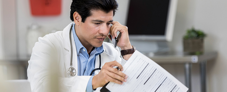 Male physician talks with colleague on the phone while reviewing a patient’s chart.