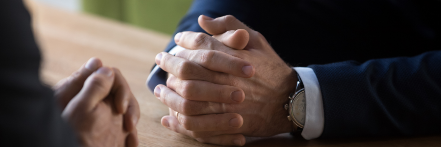 Two men facing each other at a table, each with his hands clasped together