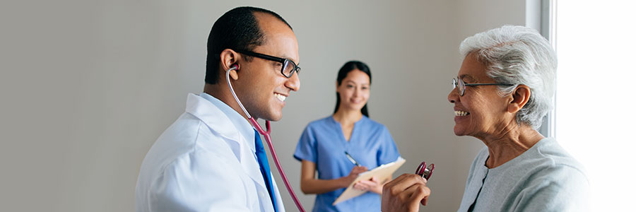 Un médecin examine une patiente tandis qu’une adjointe médicale prend des notes en arrière-plan.