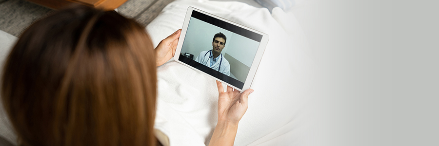 Female patient in bed holding a tablet and viewing male doctor displayed on tablet.