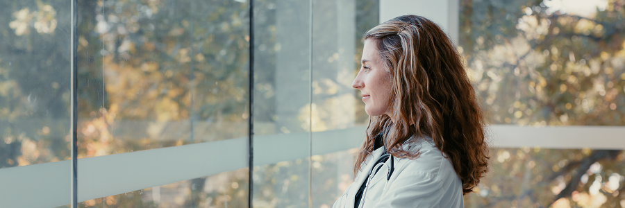 A young female doctor gazes out a large window into a natural landscape with trees, her expression is calm.