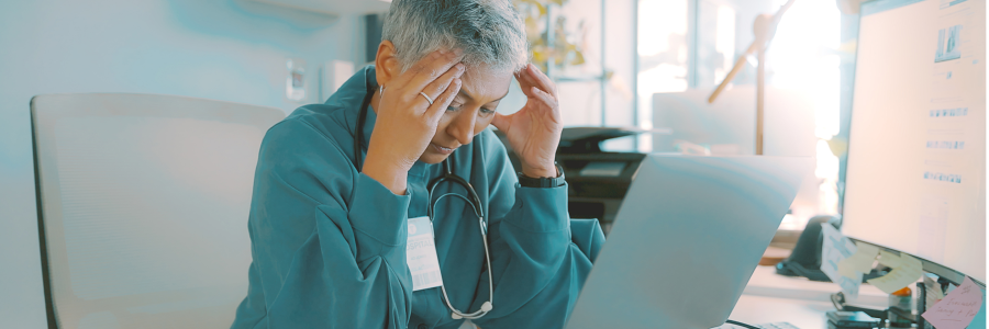 Physician at a desk looking stressed, holding their head while working on a laptop.