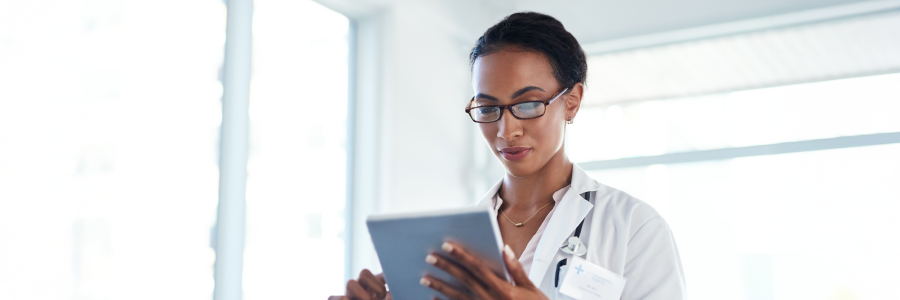 A physician in a white coat and glasses looks down at a tablet while standing in a clinical setting