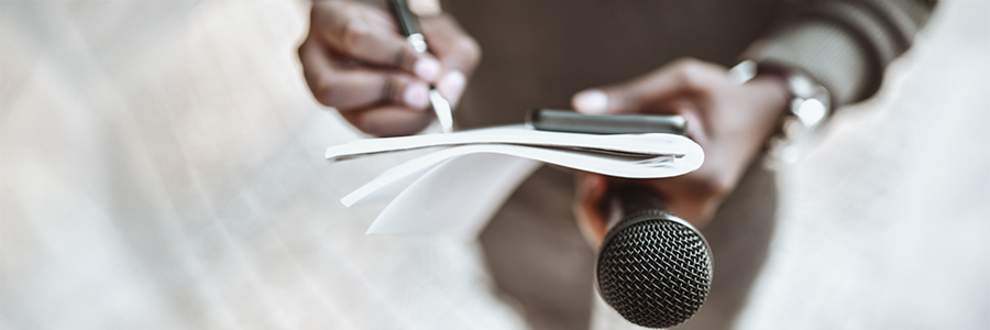 Plan sur les mains d’une personne qui tient un micro et un cellulaire tout en prenant des notes dans un carnet.