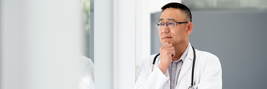 Male physician looking out a window, resting one hand on their chin in a thoughtful pose.