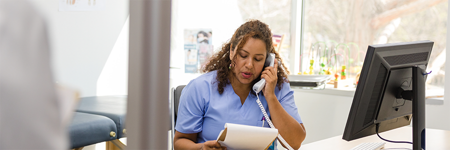 Healthcare professional seated at a computer, speaking on the phone while reviewing a paper chart.