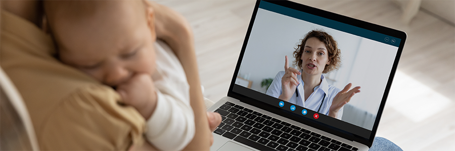 A parent holding a baby participates in a virtual medical appointment on a laptop with a physician.