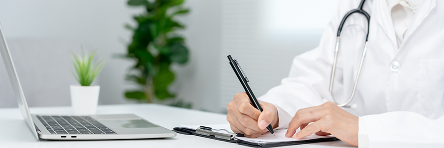 Physician seated at a desk completing a form beside an open laptop.