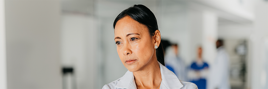 Female physician standing in a hospital setting with a pensive facial expression.
