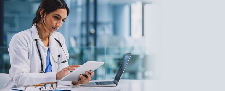 Female physician sitting at desk with a laptop and working on a tablet.