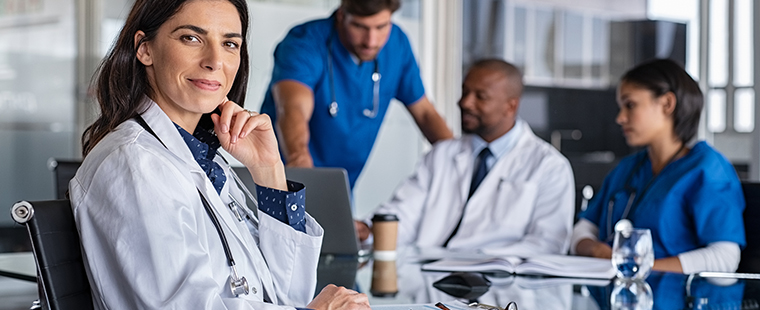 Scène de collaboration dans une salle de réunion où une médecin confiante sourit en regardant l’objectif pendant que ses collègues continuent d’échanger en arrière-plan.