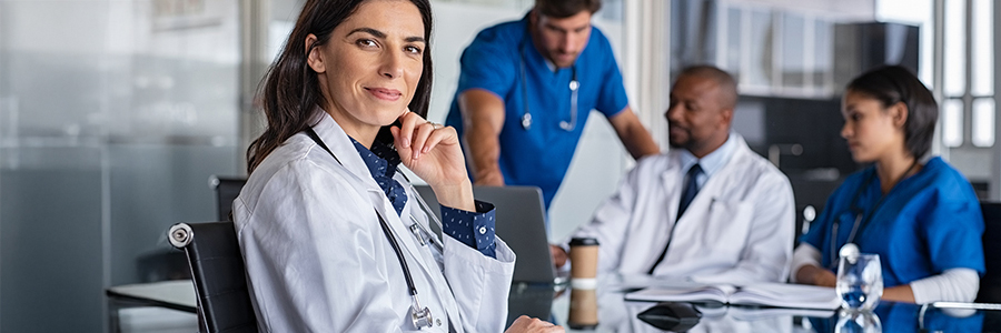 Scène de collaboration dans une salle de réunion où une médecin confiante sourit en regardant l’objectif pendant que ses collègues continuent d’échanger en arrière-plan.