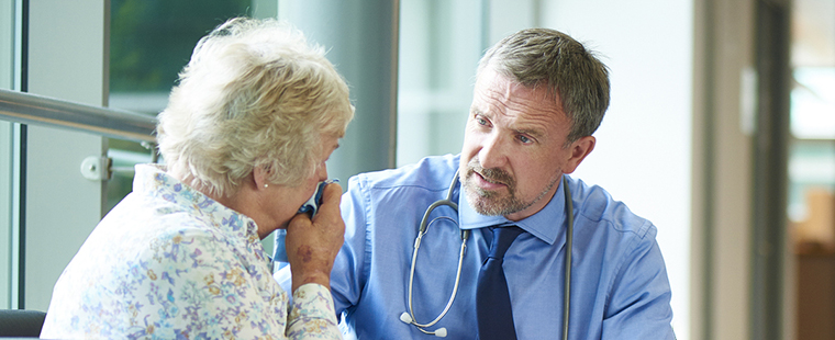 Un médecin discute avec une parente d’un patient ou une patiente bouleversée. Elle est assise dans le couloir d’un hôpital et pleure dans son mouchoir.