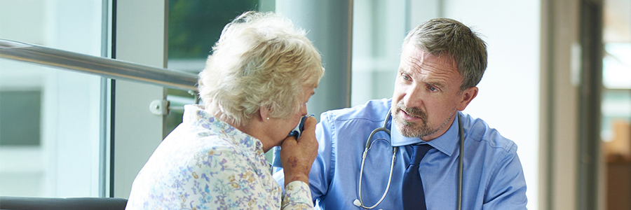 Un médecin discute avec une parente d’un patient ou une patiente bouleversée. Elle est assise dans le couloir d’un hôpital et pleure dans son mouchoir.