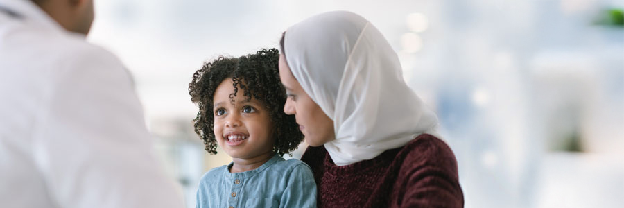 Young child sits on veiled woman’s lap and smiles at her physician. 