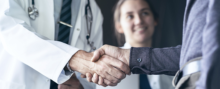 Physician and patient shaking hands with smiling resident in the background.