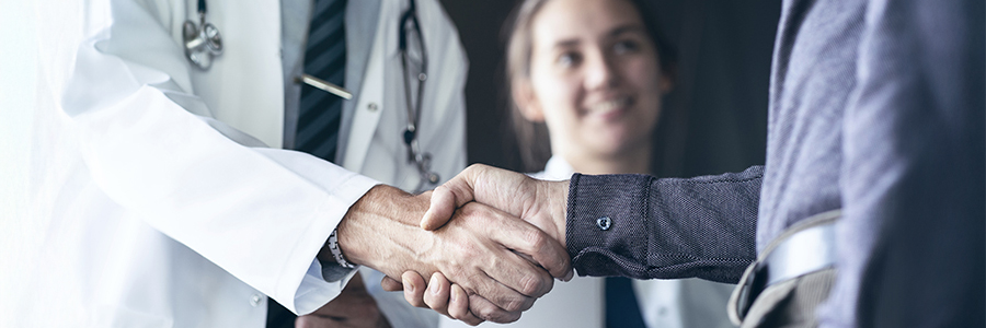 Physician and patient shaking hands with smiling resident in the background.